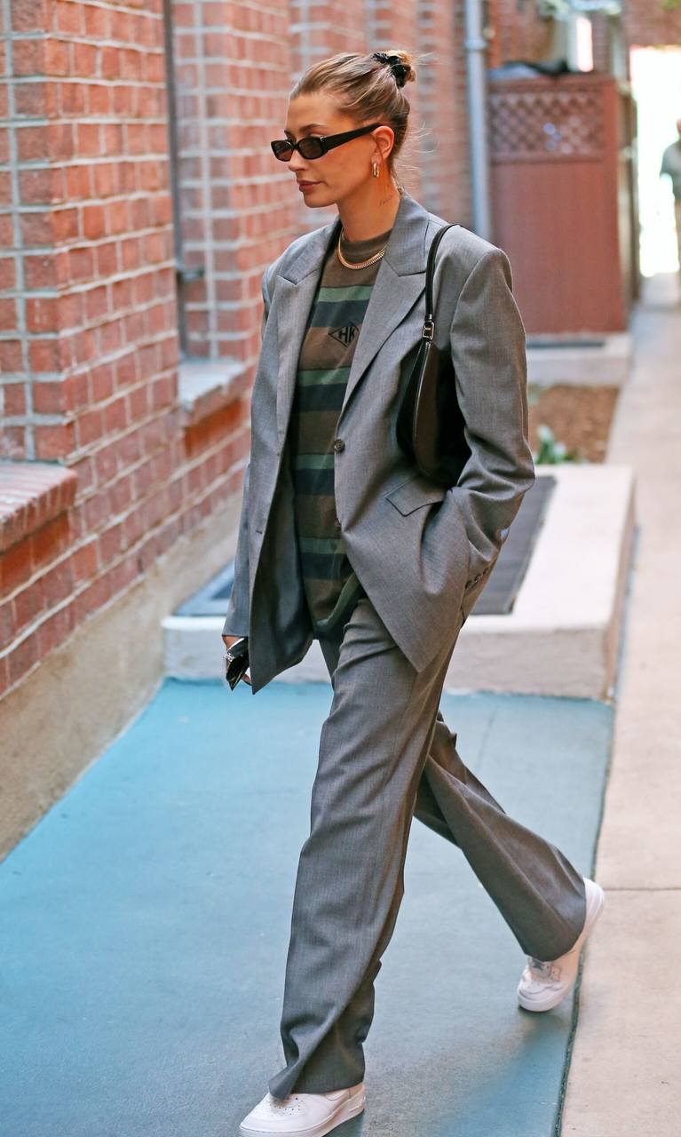 Mulher loira caminhando de óculos escuros e cabelos presos, vestida com roupa de alfaiataria, camiseta e tênis.