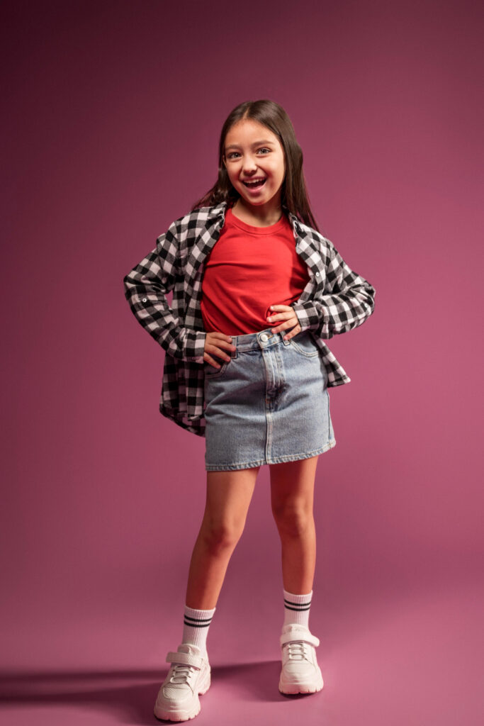 Lojistas de moda infantil: garota em fundo rosa exibindo look com tênis, meias, saia jeans, camisa xadrez preto e branca e camiseta vermelha